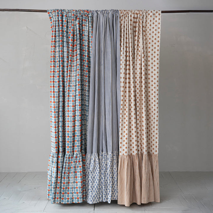 Three patterned curtain panels hanging from a dark rod against a plain light gray wall and pale floor. The left panel has a blue, white, and orange geometric pattern; the center panel is vertically striped in gray and white; the right panel has a beige background with orange geometric dots and a beige striped ruffled hem.