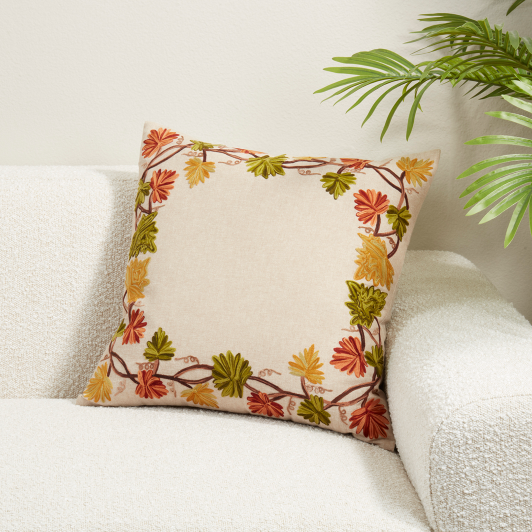 A square throw pillow with a cream center and a border of autumn leaves in orange, yellow, and green, resting on a white textured sofa with a green plant in the background.