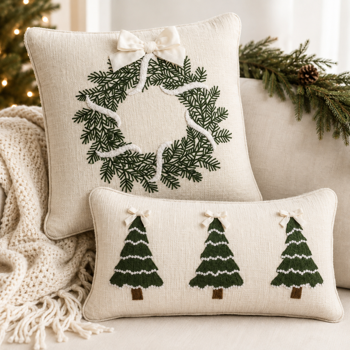 Two cream-colored linen throw pillows with embroidered green Christmas tree and pine wreath designs, styled on a sofa with a knit blanket and holiday greenery.