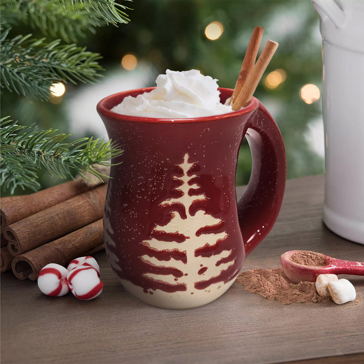 Red mug with hot chocolate, whipped cream, and cinnamon sticks on a wooden surface with Christmas decorations.