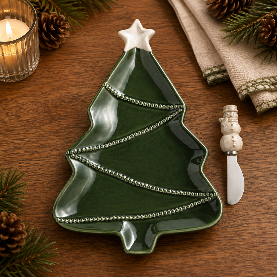 A Christmas tree-shaped green ceramic plate with silver beaded trim and a white star topper, styled on a wooden table with pinecones, a candle, a napkin, and a small snowman knife.