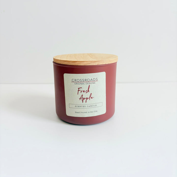 A Crossroads Original Designs scented candle in a matte burgundy jar with a light wood lid, labeled 'Fresh Apple' on a clean white background.
