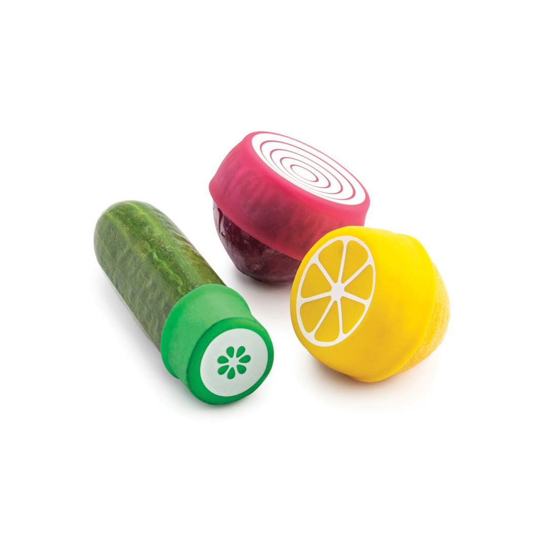 Three small vegetable-shaped containers or squeezer-like kitchen accessories on a white background: a cucumber with a green cap, a red onion half with a pink top, and a lemon half with a yellow top.