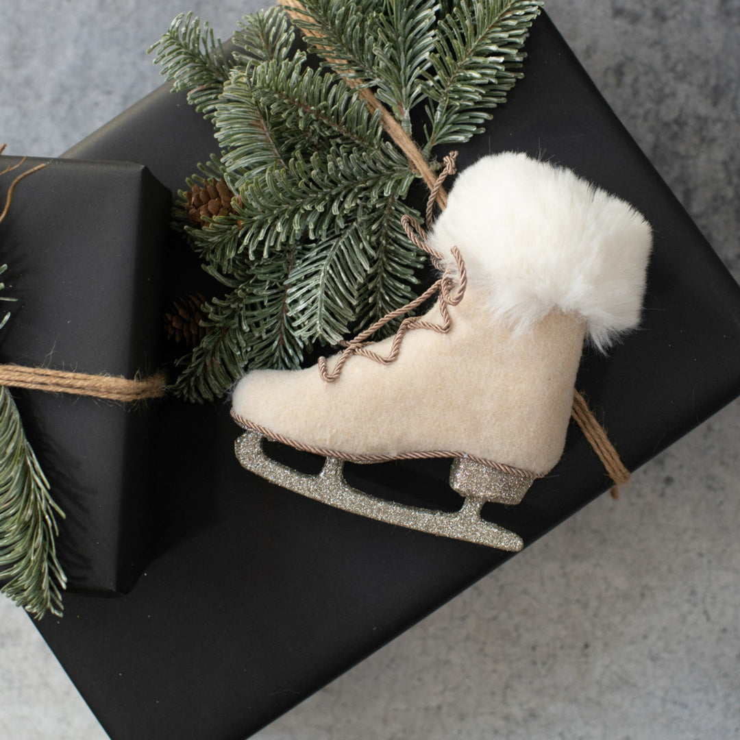 A small beige felt ice skate ornament with a fluffy white faux-fur cuff and glittery silver blade, styled on top of black wrapped gift boxes with green pine branches.