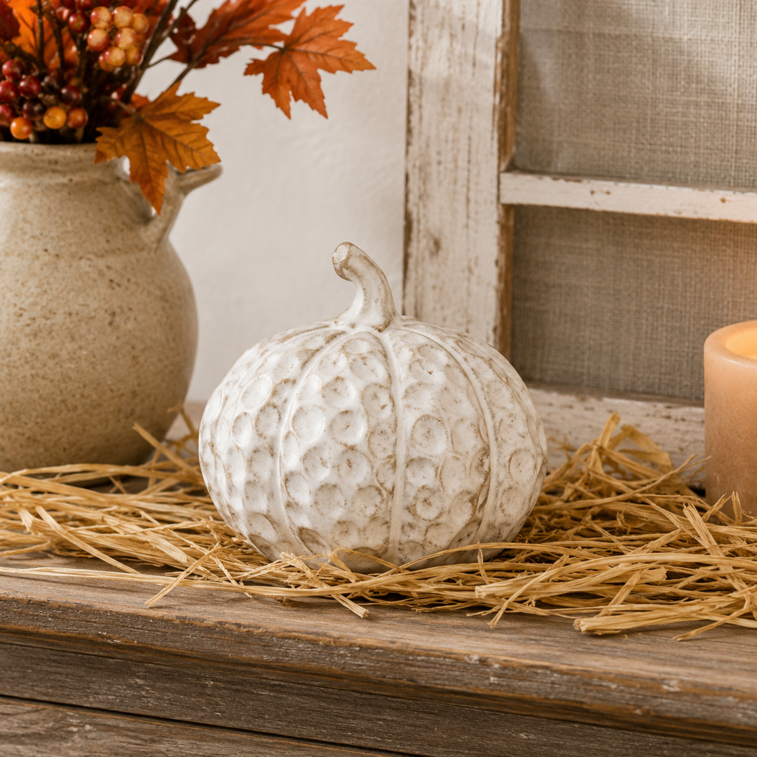 A white textured decorative pumpkin sits on a bed of straw on a rustic wooden surface, with a beige ceramic vase of autumn leaves and a lit candle in the background.