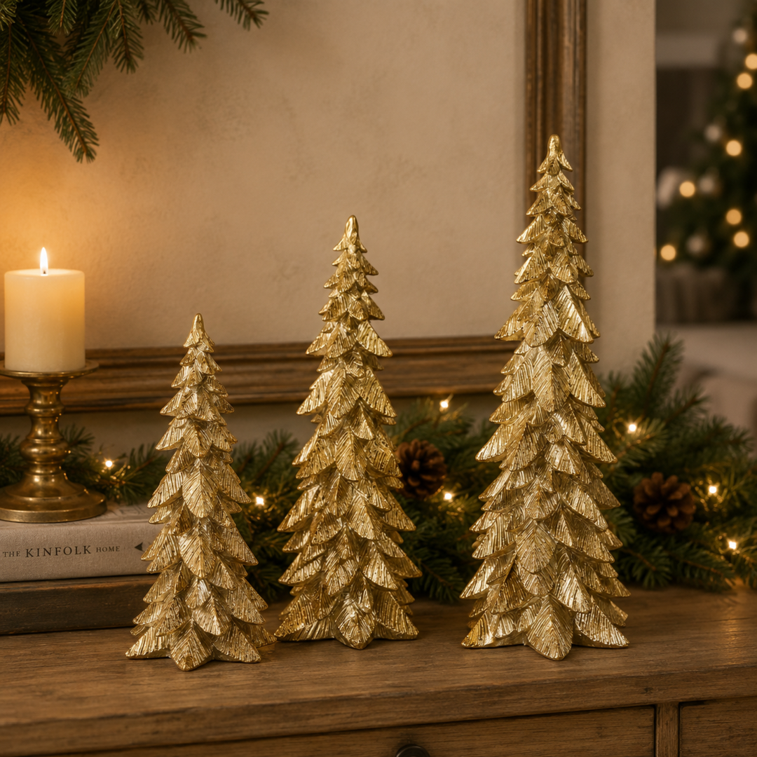 Three small metallic gold Christmas tree ornaments of varying heights displayed on a wooden table, surrounded by green pine garlands, lit candles, and warm holiday lights.