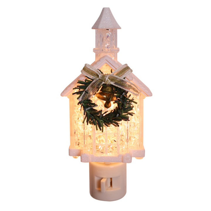 A Christmas birdhouse-shaped night light with warm white LED lights inside, decorated with a green pine wreath, gold bell, and a glittery gold ribbon bow on a white plastic base.