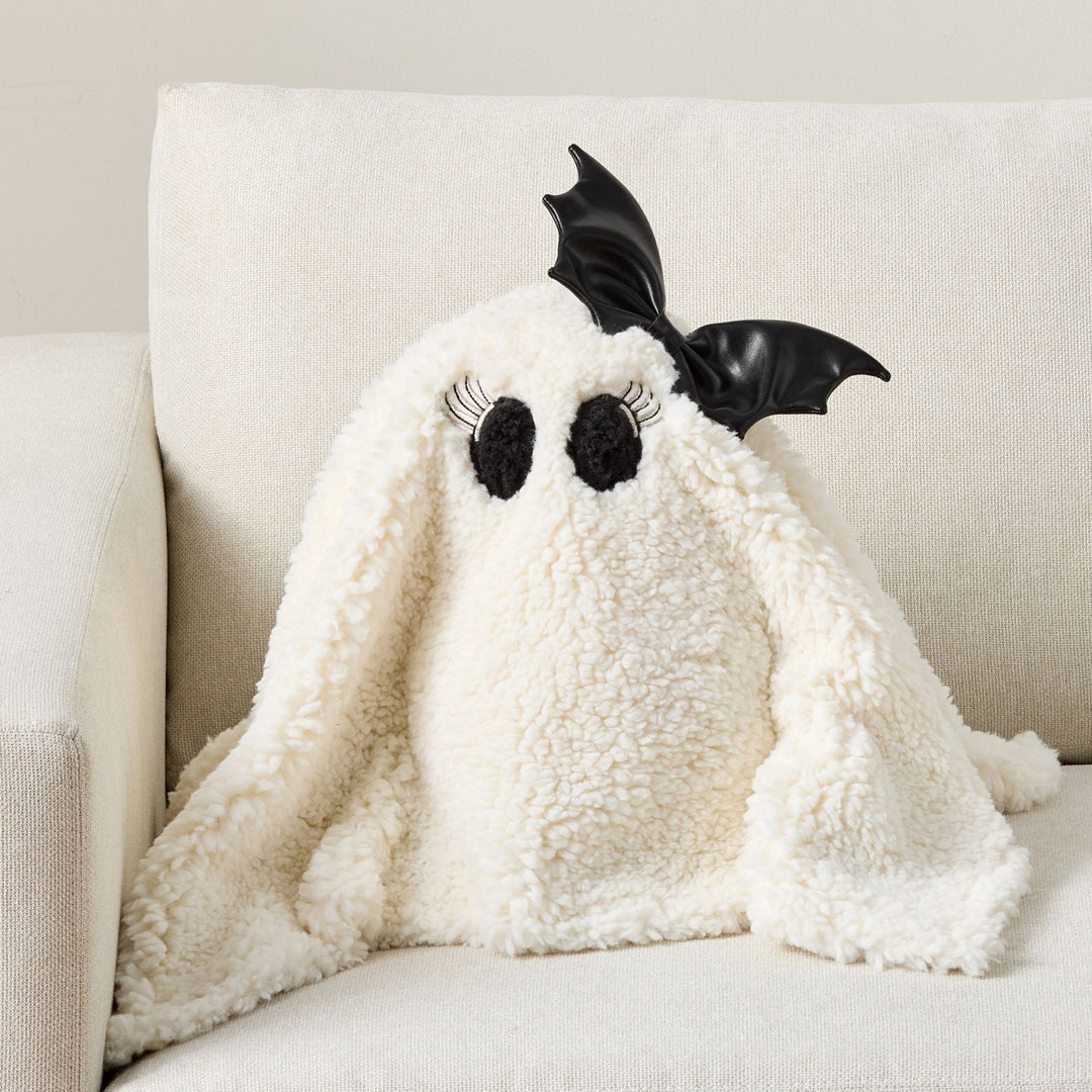 A plush ghost-shaped baby blanket or pillow in textured cream fleece, styled on a light beige upholstered armchair. The ghost has black bat wings, black embroidered eyes with white eyelashes, and a black nose.