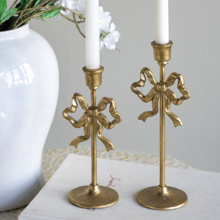 Two gold candlestick holders with bow designs on a white surface.