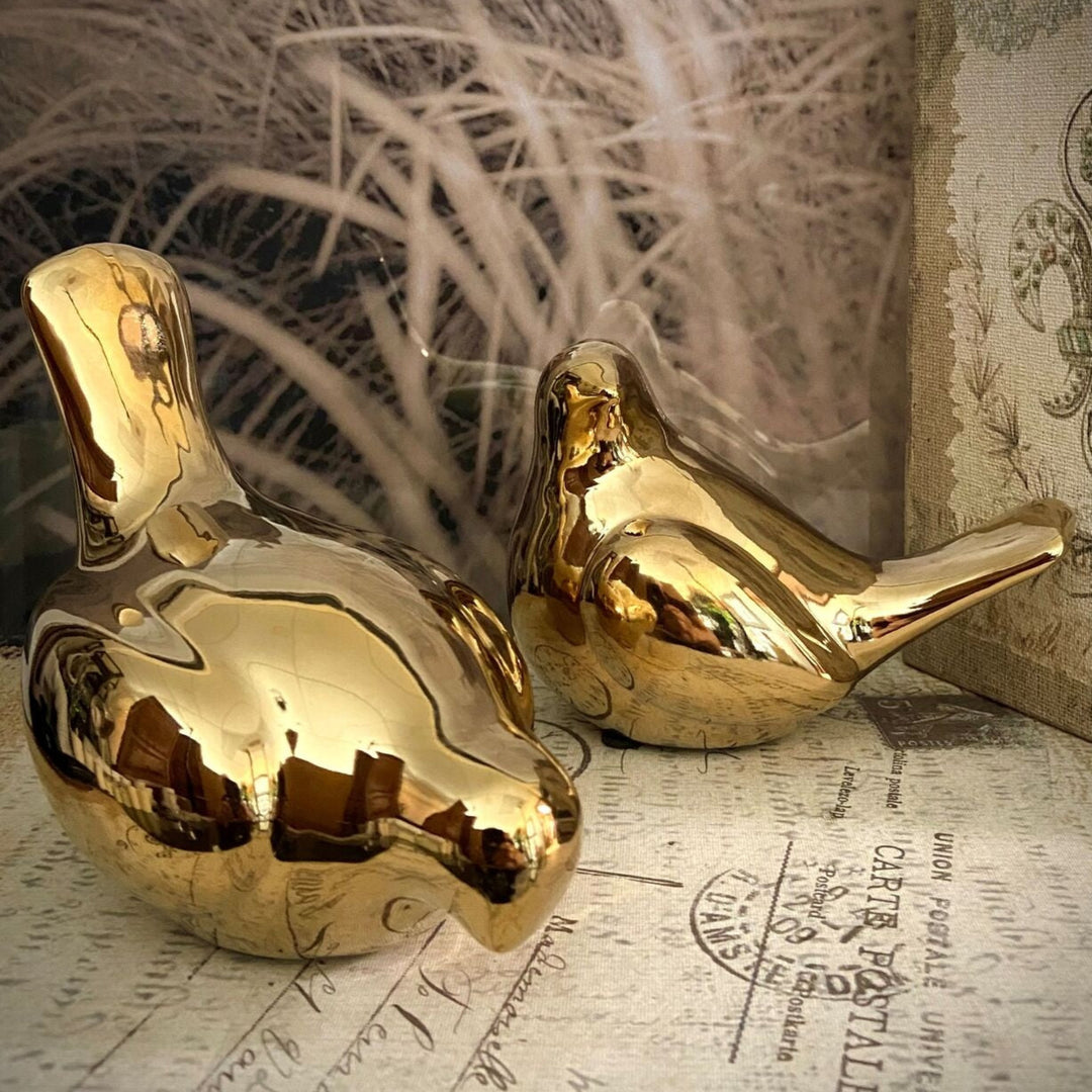 Two shiny gold decorative bird figurines resting on vintage-style printed paper with soft beige feathers in the background.