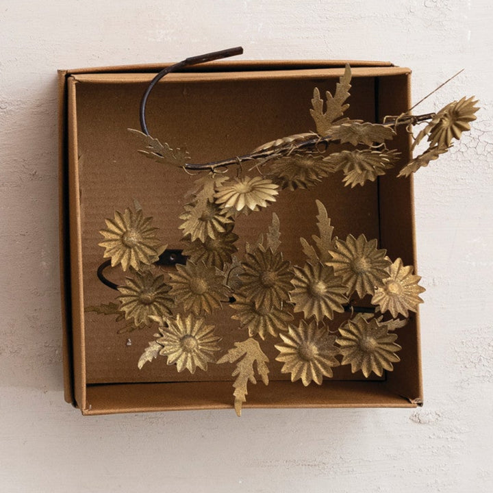 Three open brown cardboard hair clips are arranged on a light textured background. One clip holds a green leafy branch, one holds a cluster of pink and white daisy-like flowers with green leaves, and one holds a cluster of metallic gold leafy branches.