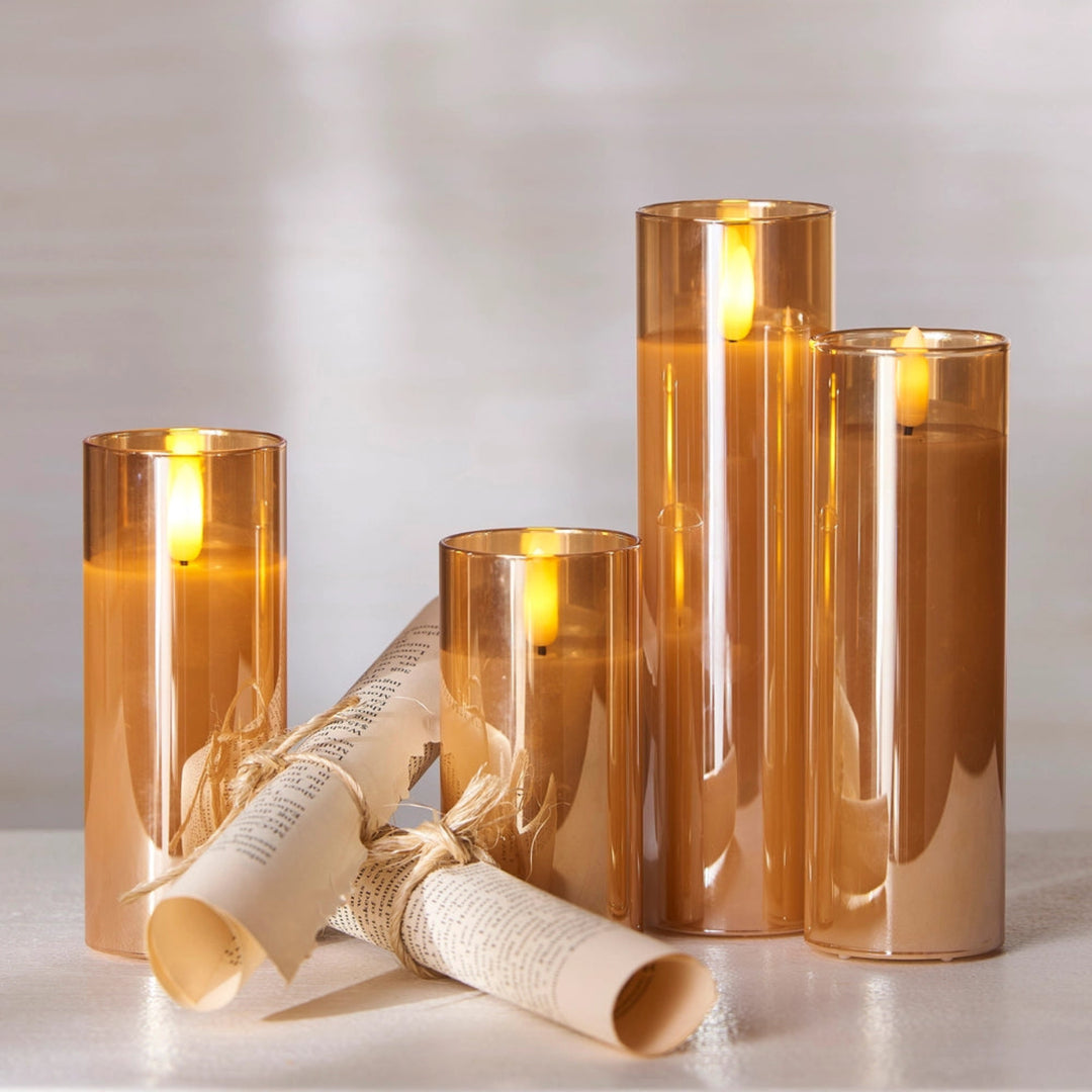 Four amber-tinted glass pillar candles of varying heights, some lit with warm yellow flames, arranged on a light surface with two rolled parchment-style accents in the foreground.