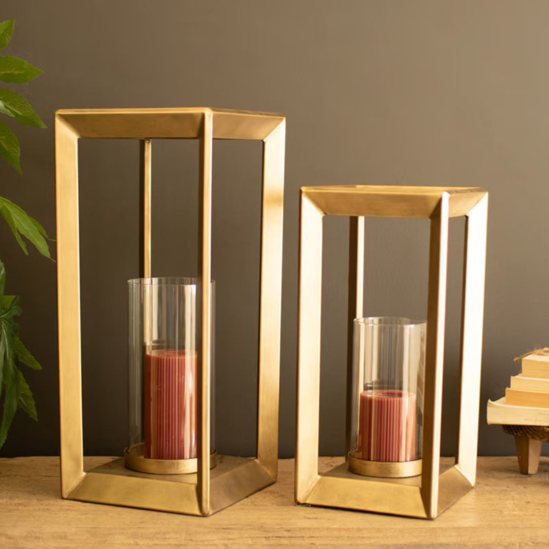 Two gold-toned metal lantern candle holders with clear glass cylinders and pink candles, displayed on a wooden surface against a dark gray wall with a plant and books as props.