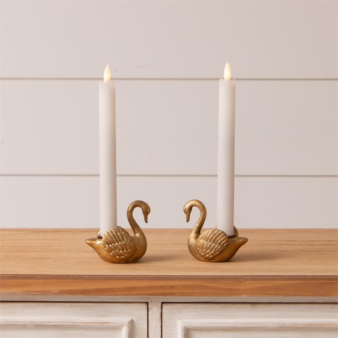 Two gold swan-shaped taper candle holders with candles on a wooden surface