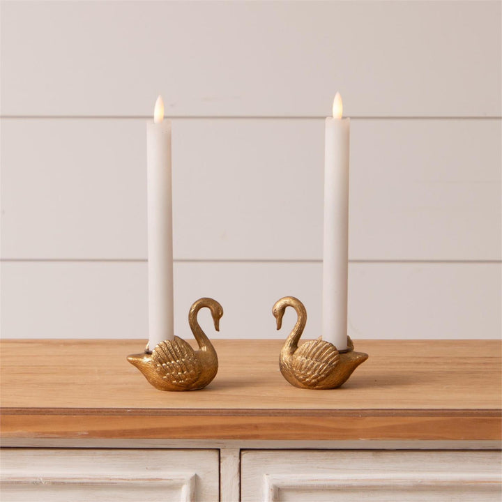Two gold swan-shaped taper candle holders with candles on a wooden surface