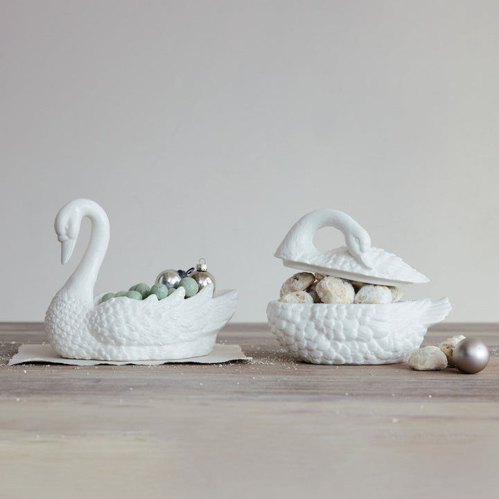 Two white swan-shaped ceramic bowls on a wooden surface with decorative items inside.