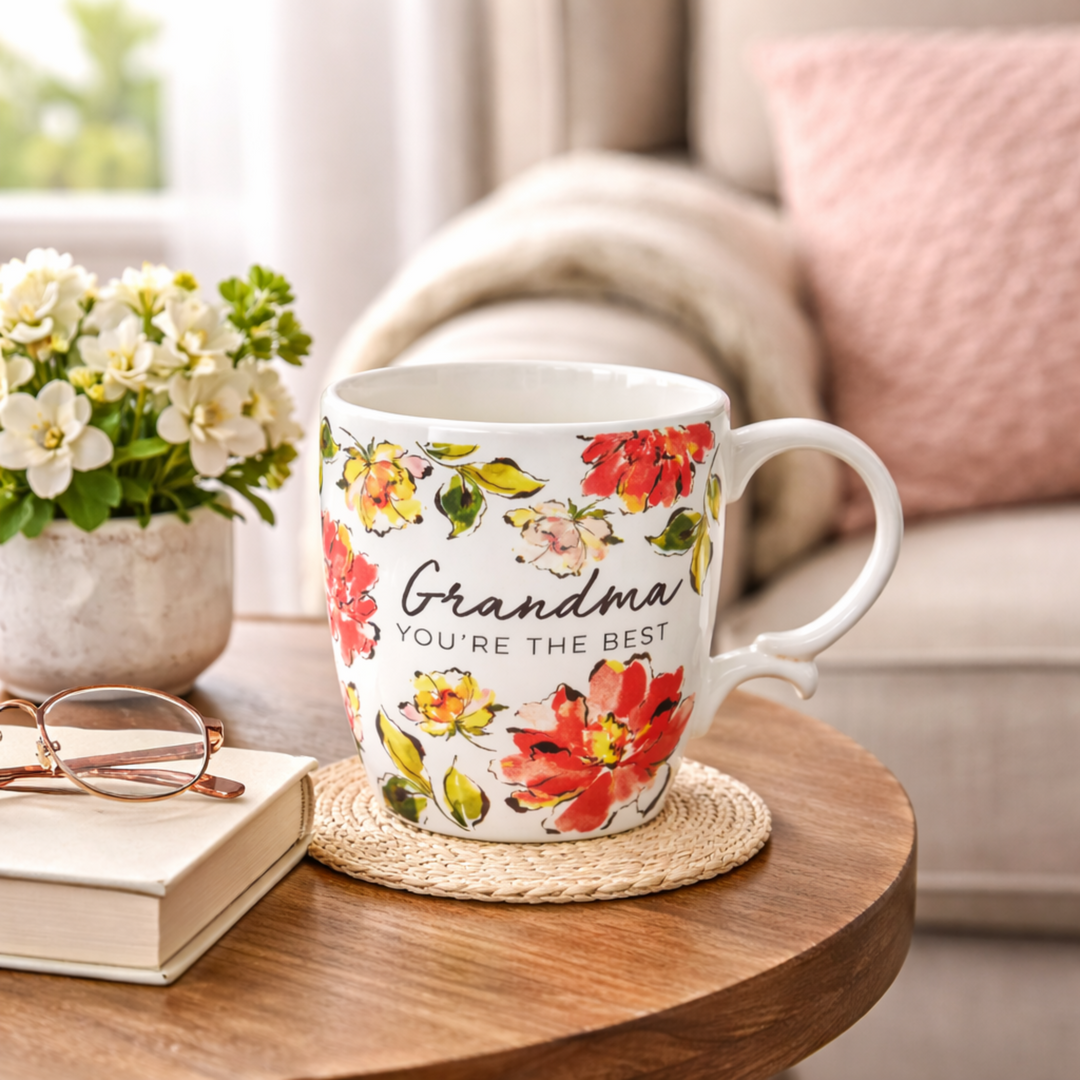 Floral mug with 'Grandma, you're the best' text on a wooden table with a book and glasses.
