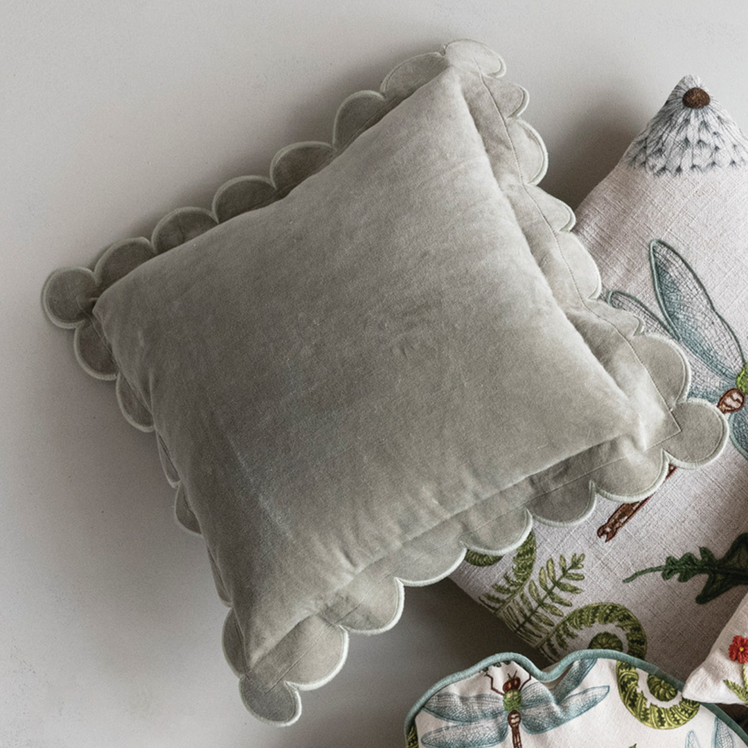 A square light gray velvet pillow with scalloped edges, styled on top of a white linen pillow with green dragonfly and fern embroidery.