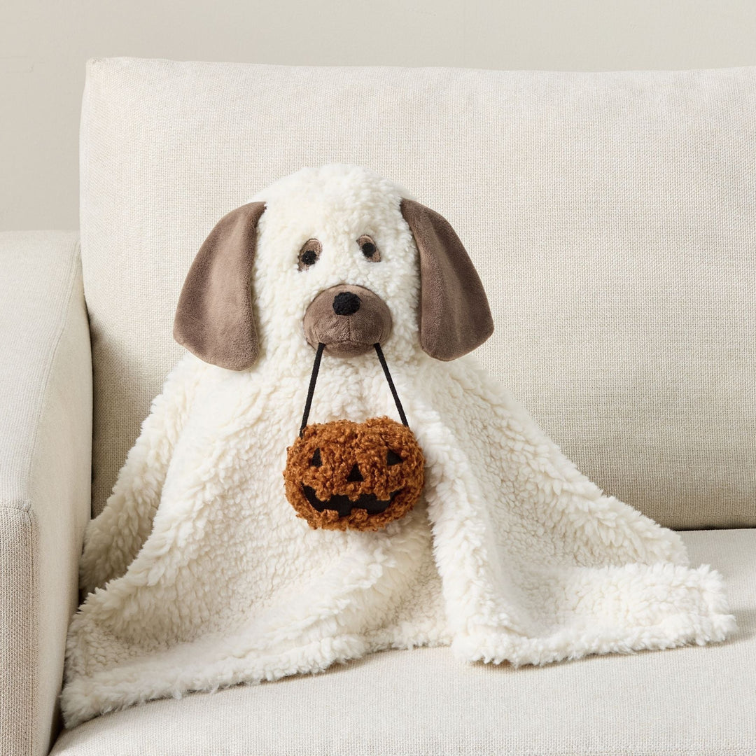 A plush dog-shaped throw blanket in off-white faux fur with brown ears, nose, and paws, sitting on a cream-colored sofa and holding a small brown crocheted pumpkin bag.