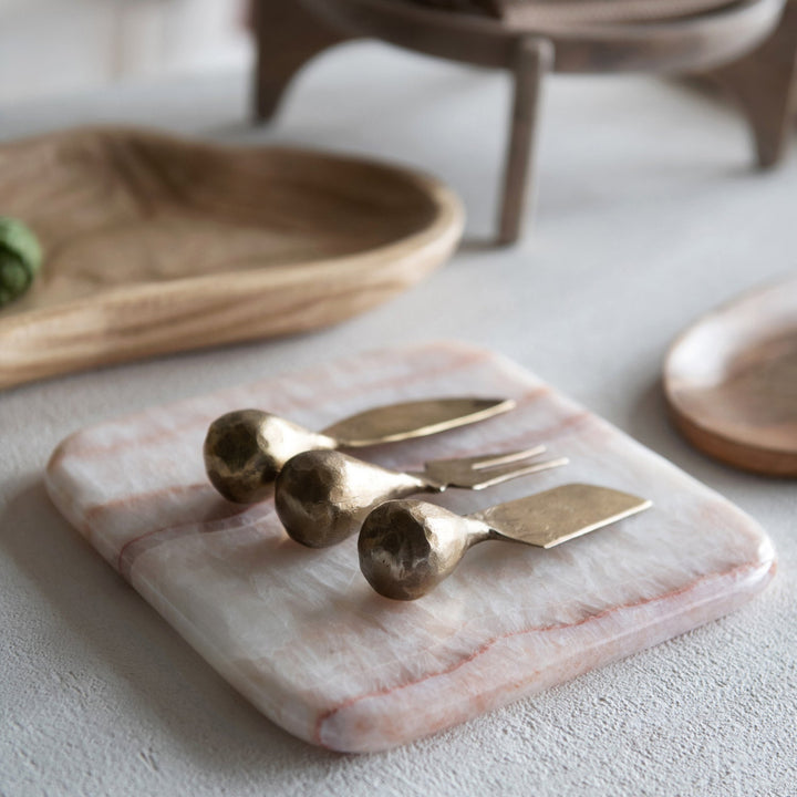 Set of brass cheese knives on a pink marble board with wooden plates in the background.