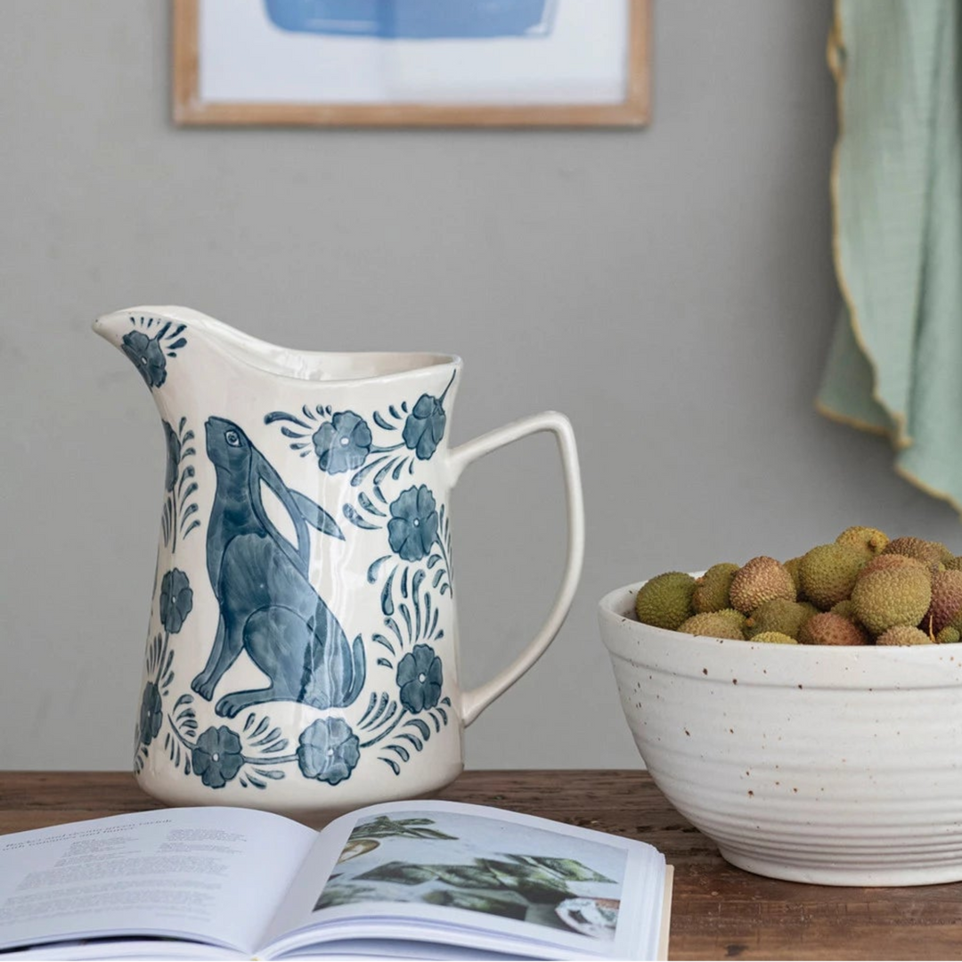 Hand Painted Rabbit & Flowers Stoneware Pitcher on a wooden table in front of a book. bowl of fruit to the side with a tea towel and photo frame in the background