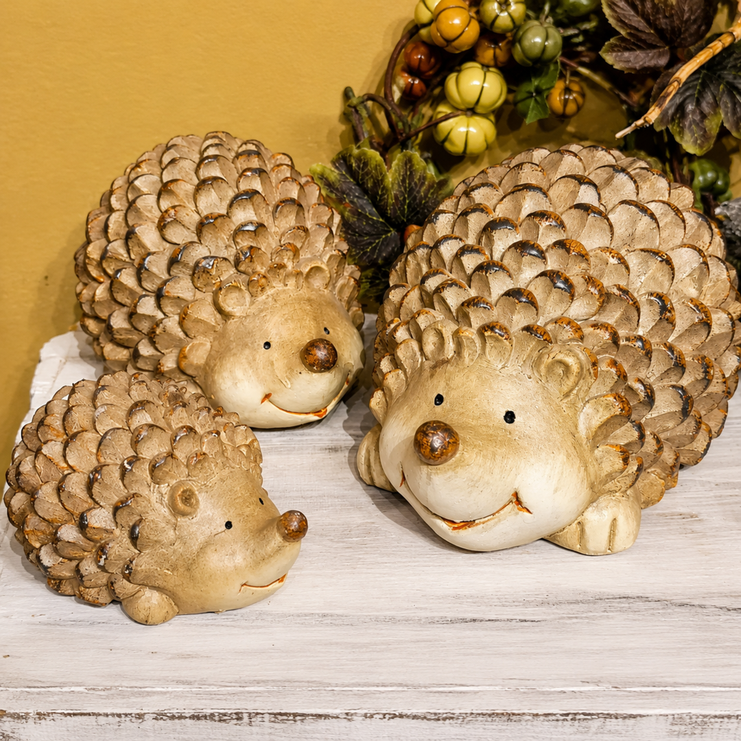 Three ceramic hedgehog figurines in different sizes, styled on a white wooden surface with a yellow background and autumn foliage.