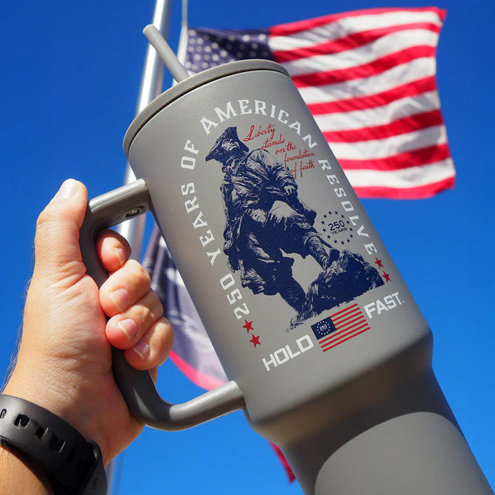 Gray mug with American Resolve design held by a hand against a blue sky with an American flag.