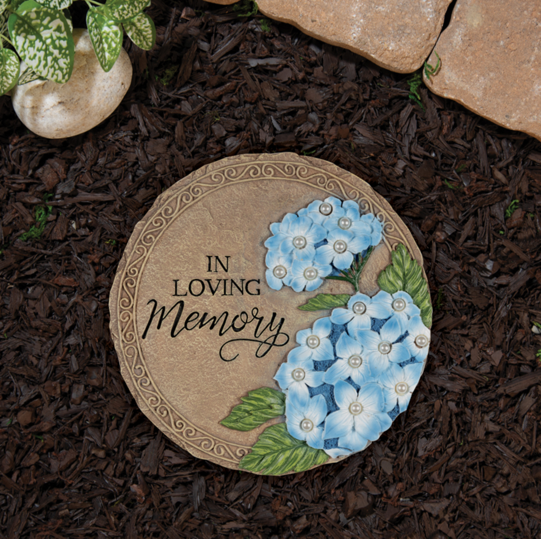 Round decorative stone with blue flowers and 'In Loving Memory' text on a garden setting.