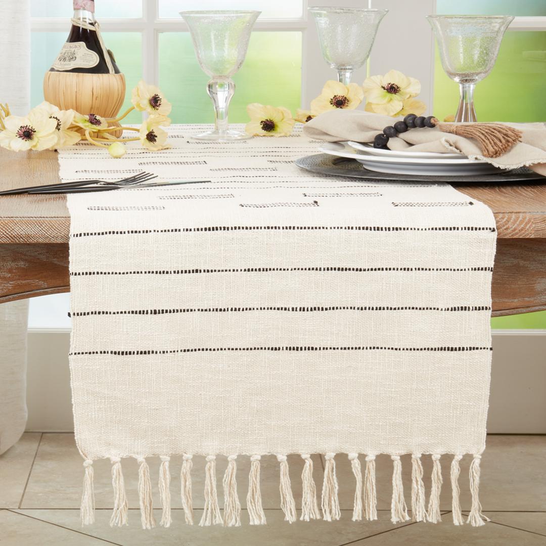 Table setting with a white tablecloth featuring black lines and fringes, accompanied by wine glasses and flowers.