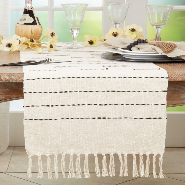 Table setting with a white tablecloth featuring black lines and fringes, accompanied by wine glasses and flowers.