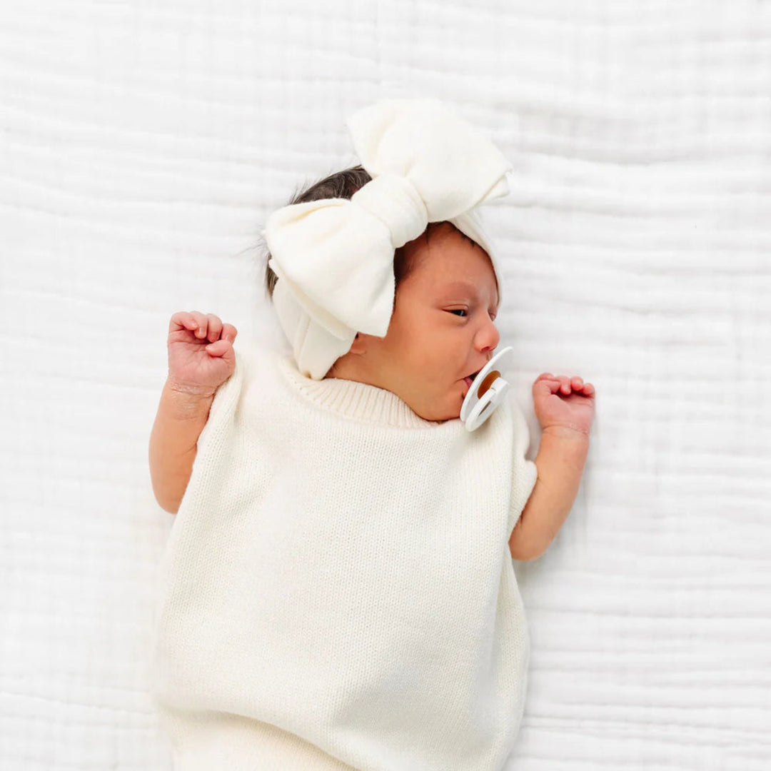A newborn baby lying on a white textured blanket, wearing a cream-colored knit swaddle blanket and a matching cream bow headband, with a pacifier in their mouth.