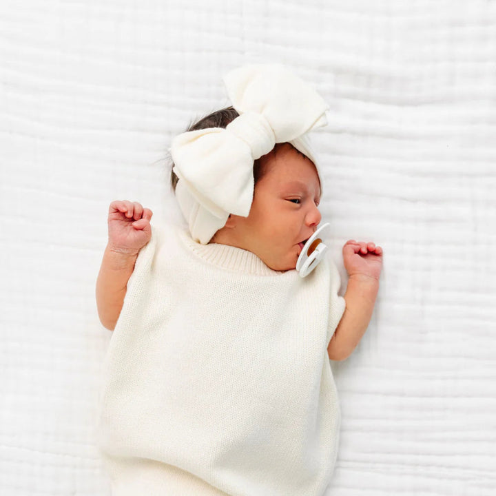 A newborn baby lying on a white textured blanket, wearing a cream-colored knit swaddle blanket and a matching cream bow headband, with a pacifier in their mouth.