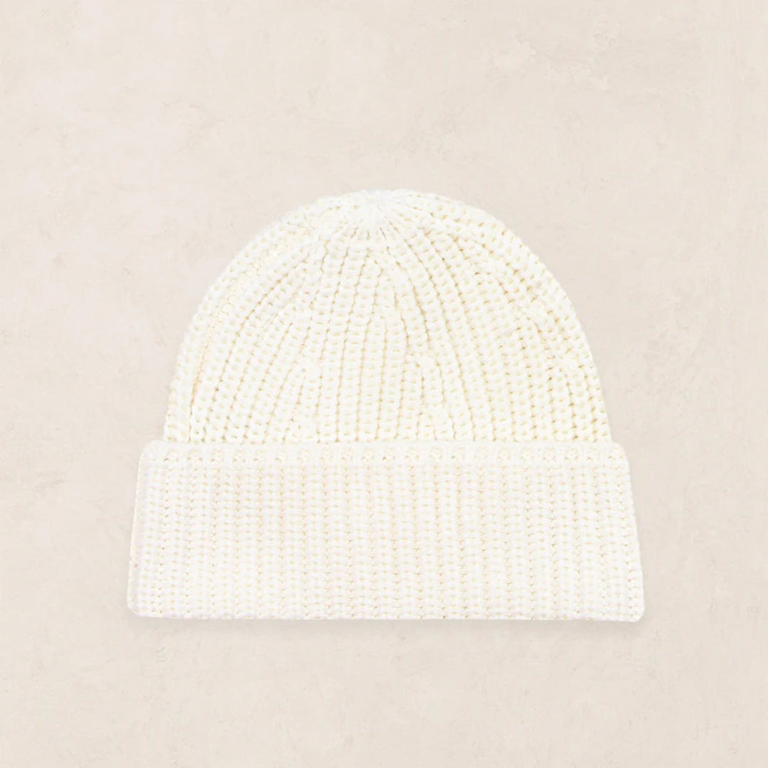 An off-white ribbed knit beanie hat with a folded cuff, displayed flat against a textured beige background.