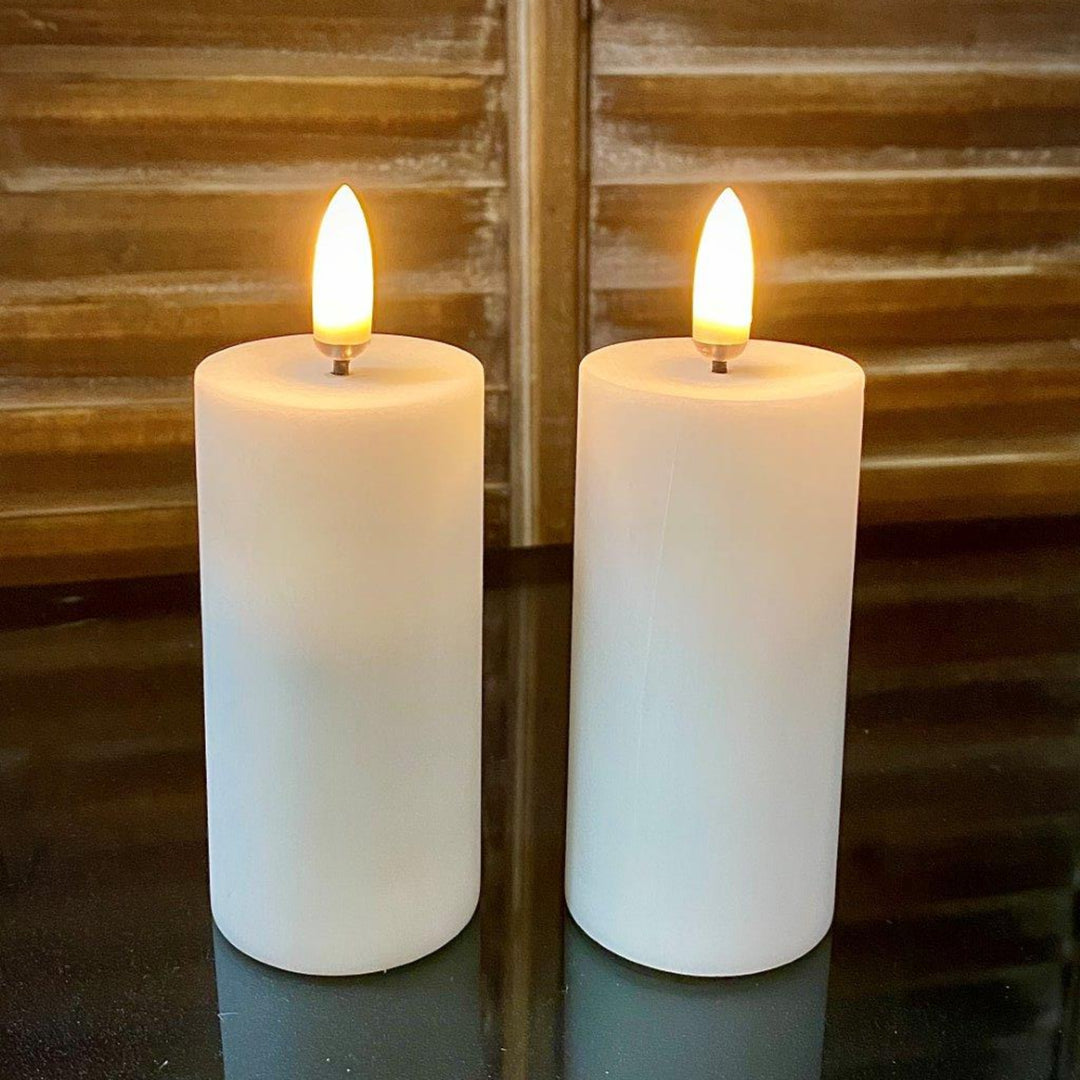 Two white candles with lit wicks on a reflective surface with a wooden background