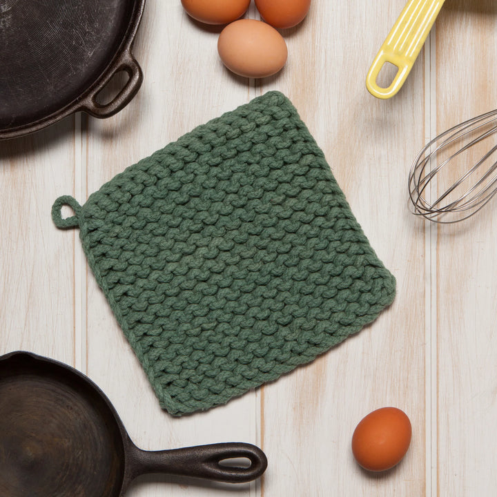 Green knitted pot holder on a wooden surface with eggs and kitchen tools.