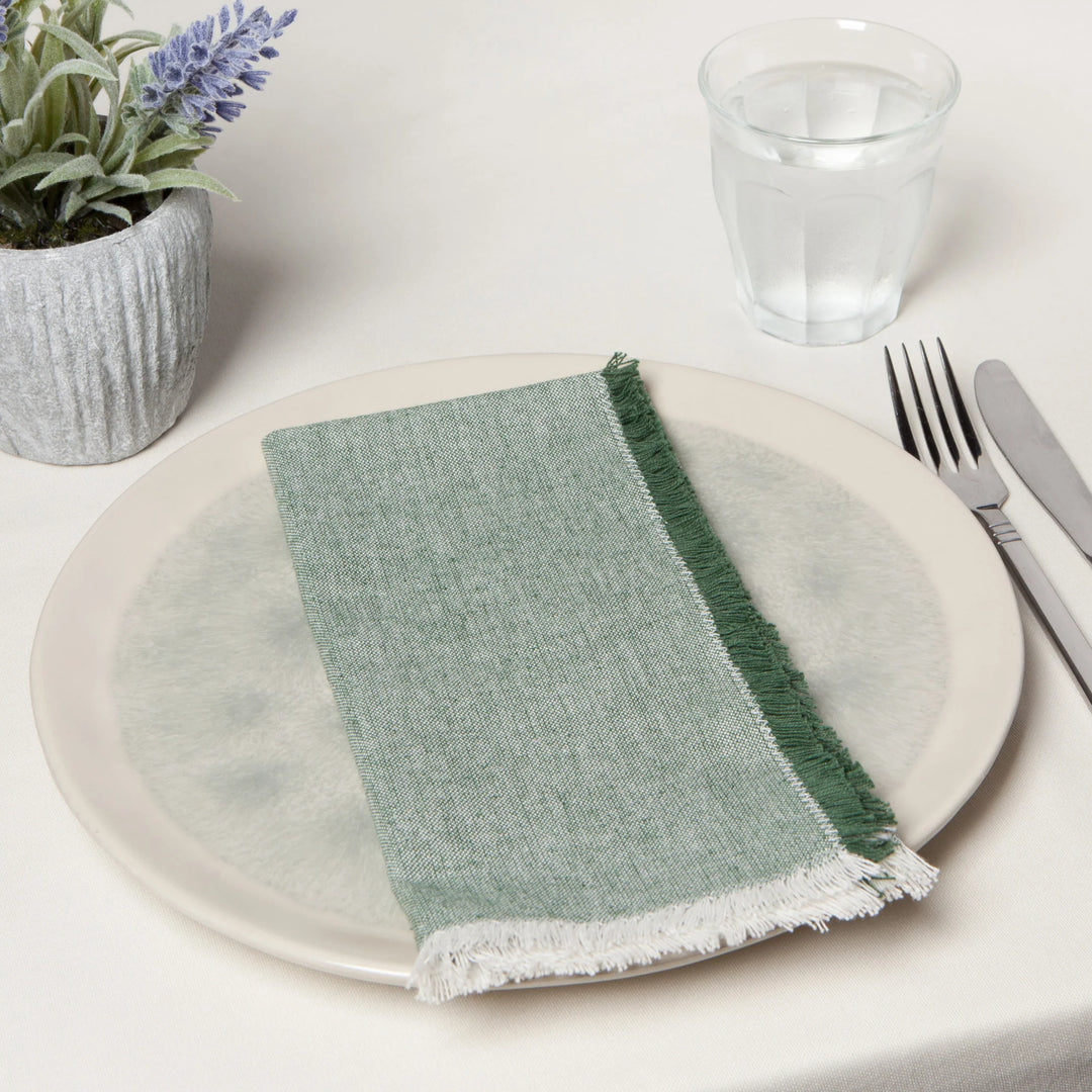 Green napkin on a white plate with a glass of water and cutlery on a white surface.