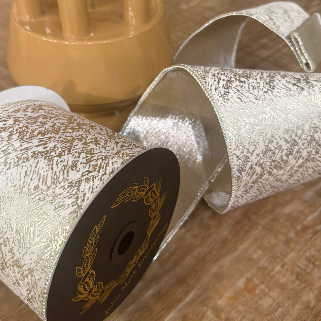 A close-up of a spool of textured glitter ribbon with a metallic silver body and gold foil flecks, shown on a wooden surface with a blurred decorative object in the background.