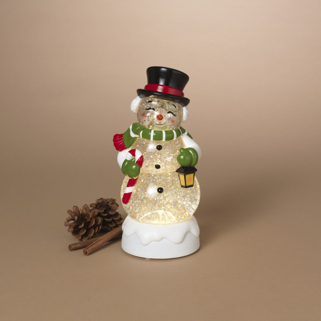 A snowman figurine with a glittery gold body, wearing a black top hat with a red band, a green and white striped scarf, green mittens, and holding a red-and-white candy cane and a small lantern. It stands on a white base against a plain beige background, with a pinecone and cinnamon stick beside it.