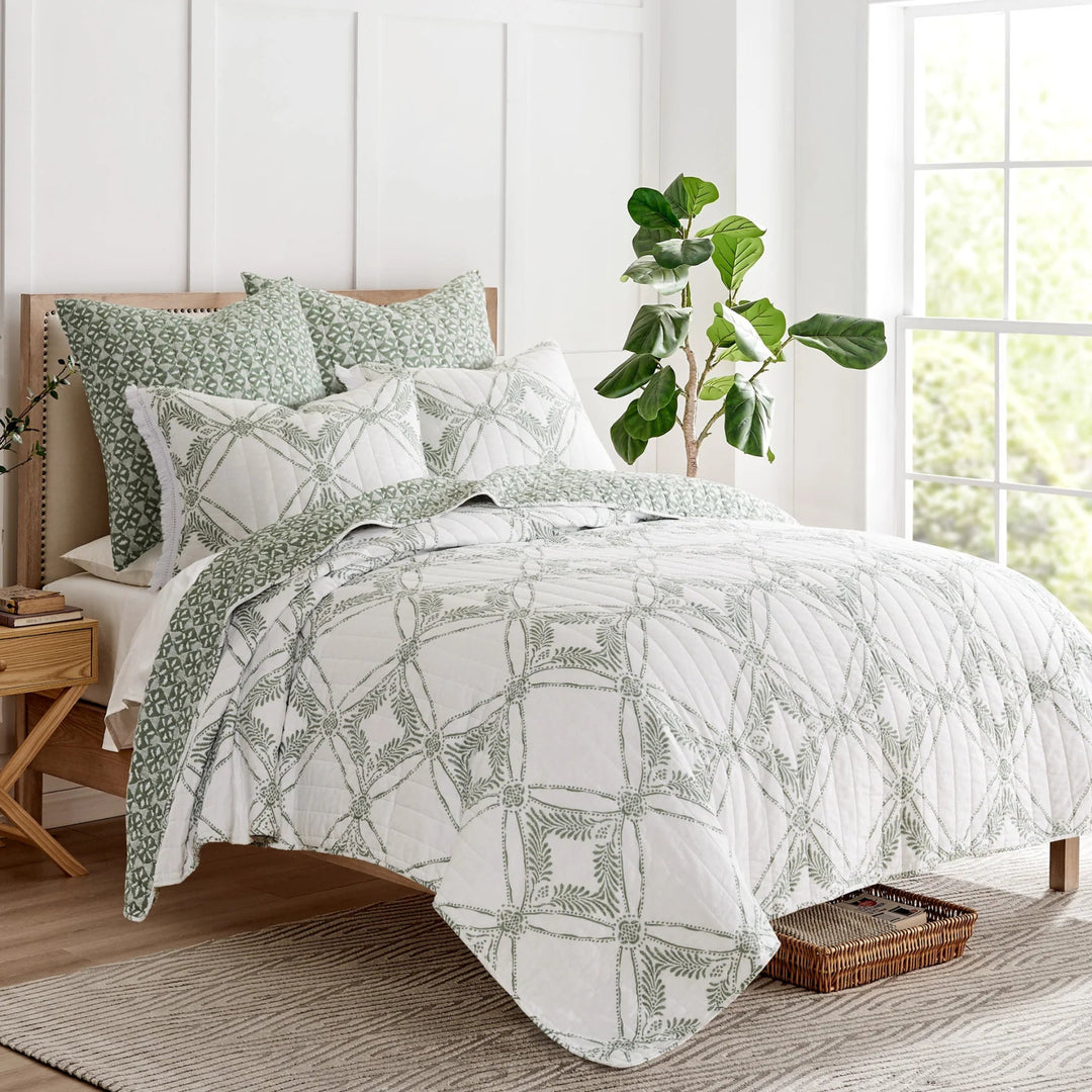 Bedroom with green and white patterned bedding, plant, and wooden nightstand.
