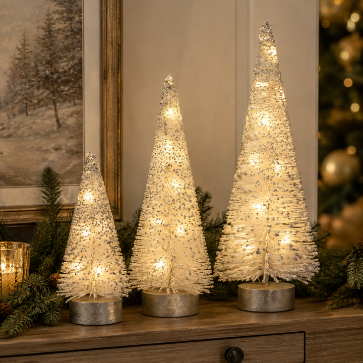 Three small frosted white Christmas trees with warm white string lights and silver bases, displayed on a wooden surface with holiday greenery and a softly blurred Christmas tree in the background.