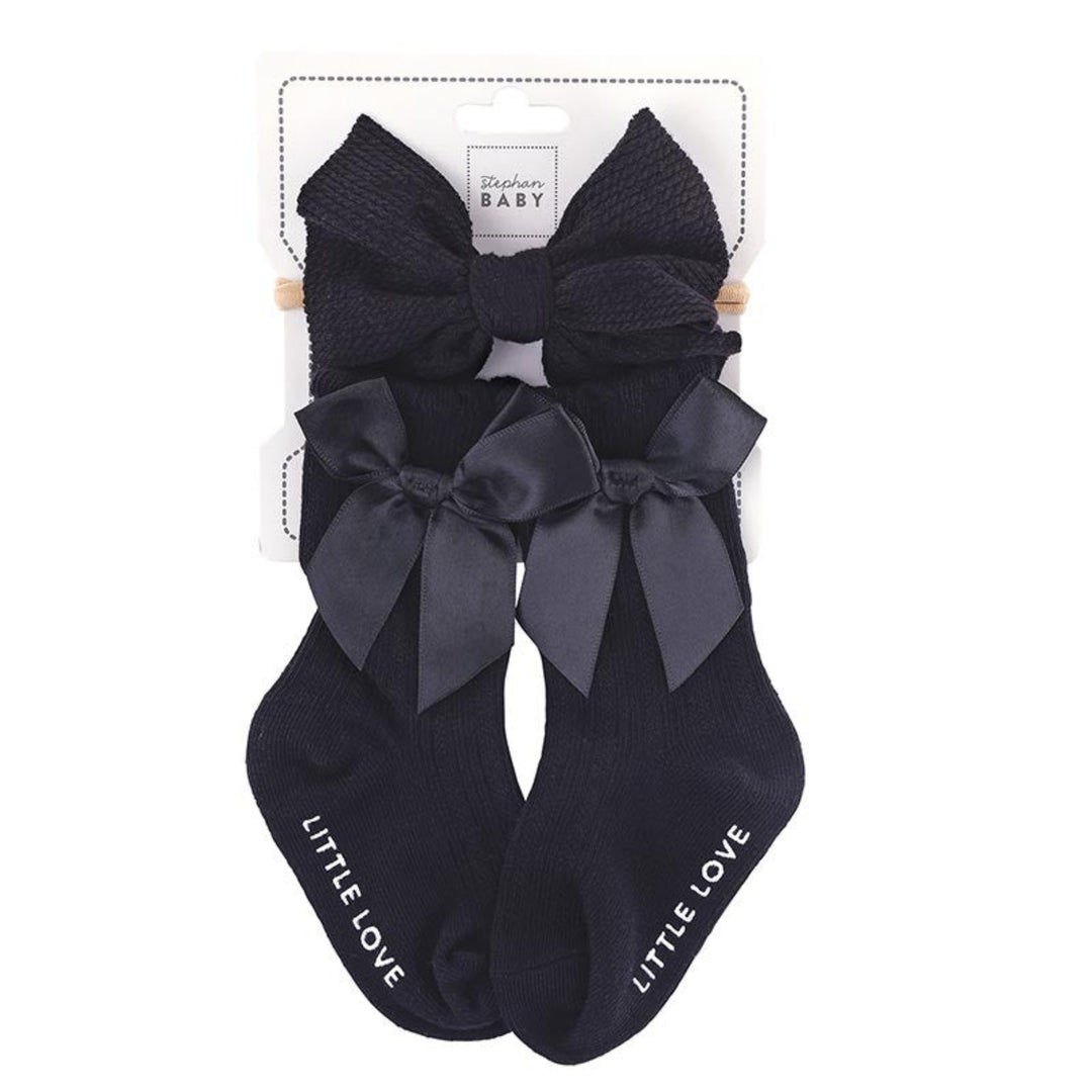 A pair of black baby socks with large matching bows on the toes, packaged on a white card labeled 'St. Lukes BABY'.