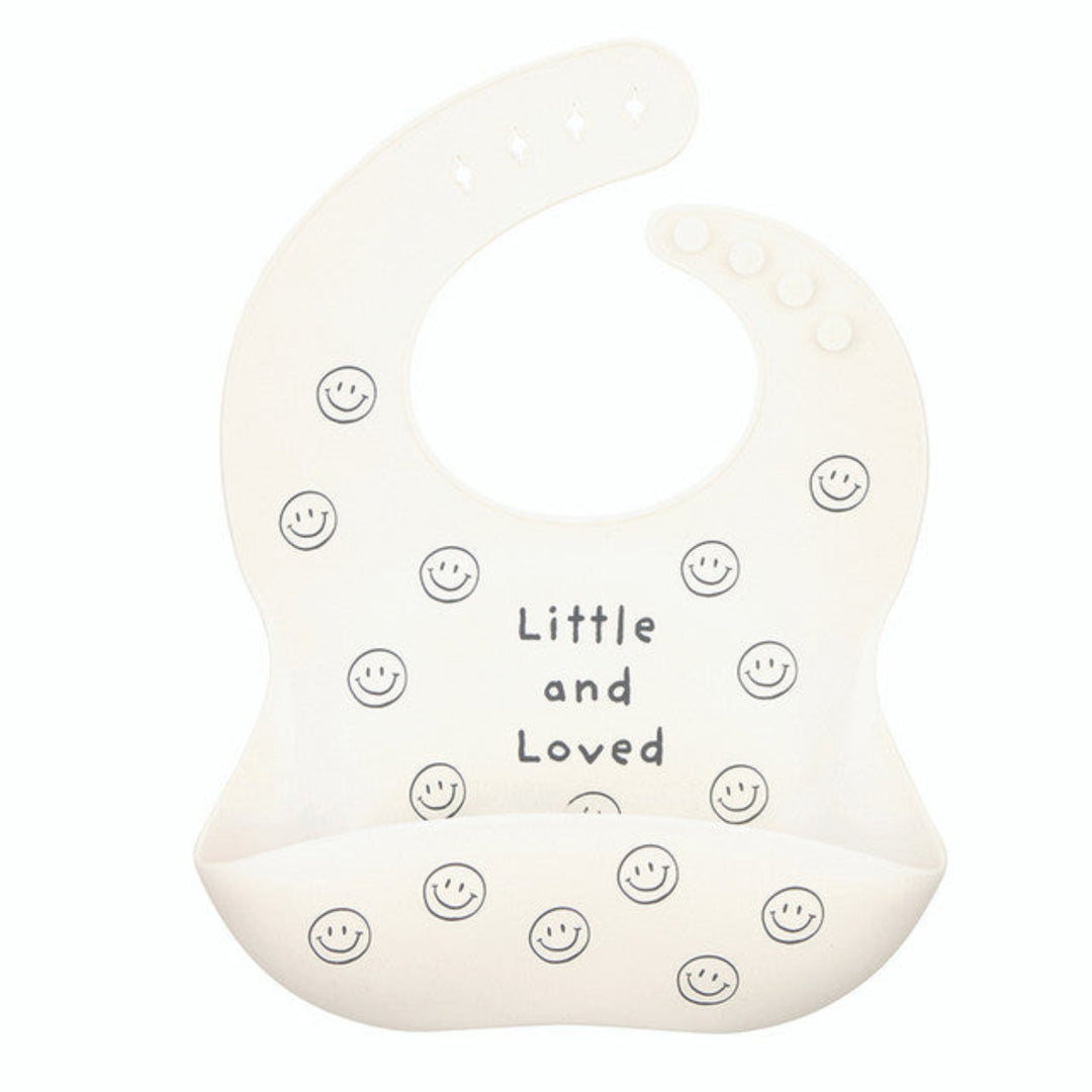 An off-white silicone baby bib with a printed design of small smiley faces and the text 'Little and Loved' in the center, set against a plain white background.