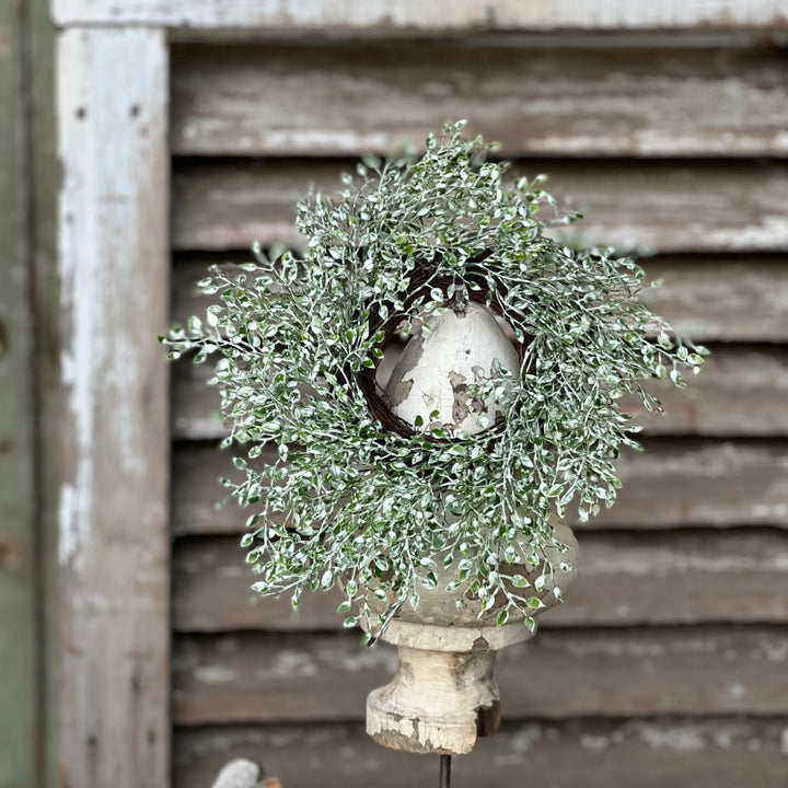 A green candle ring wreath with a leafy texture displayed on a white surface, against a wooden backdrop.