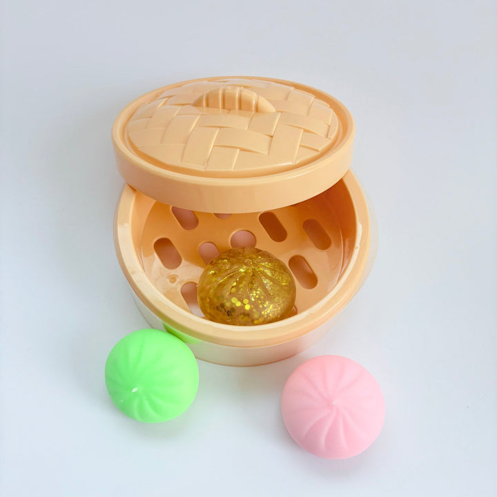 A beige plastic toy steamer with a woven-pattern lid, shown open with a gold foil ball inside and two small round balls in green and pink placed in front, on a light gray background.