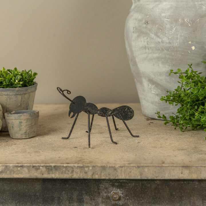 Decorative metal ant on a wooden surface with plants and a vase in the background