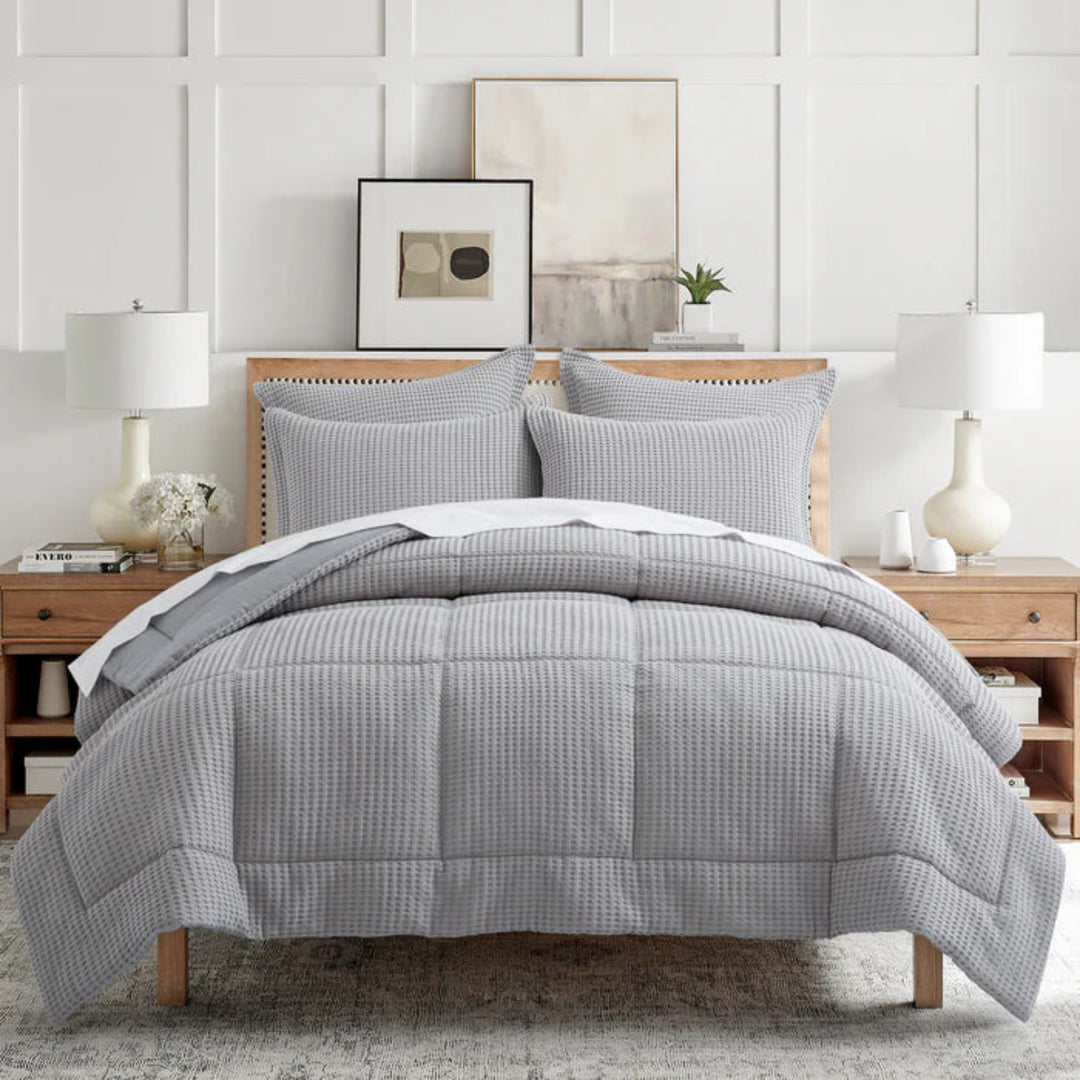 A neatly made bed in a bright, modern bedroom setting, featuring a gray and white waffle-textured comforter set with matching pillow shams, wooden nightstands, and white table lamps.