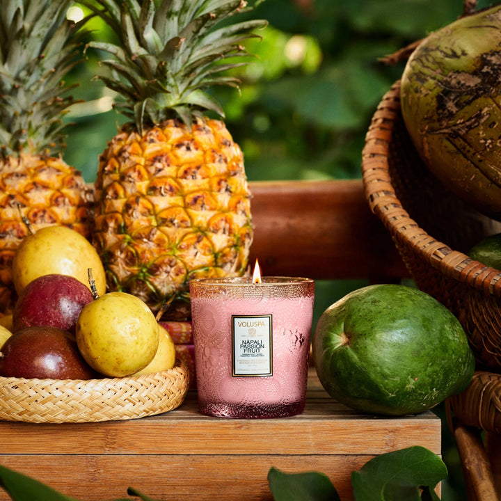 Candle with 'Napali Fruit' label surrounded by pineapples, fruits, and a wicker basket on a wooden surface.