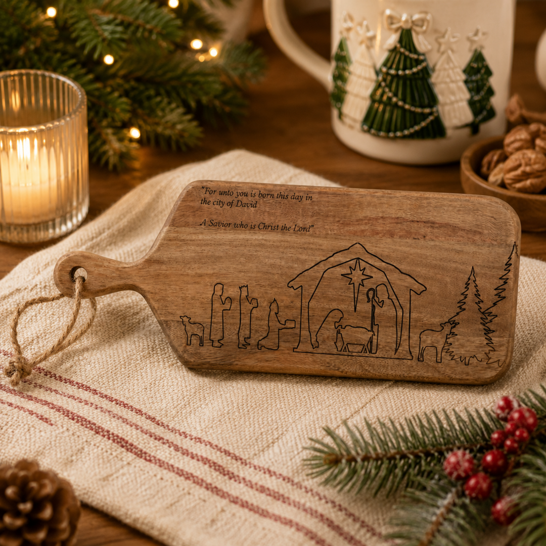A wooden cutting board with a nativity scene engraving and a hanging loop, styled on a striped linen cloth with a lit candle, pine branches, and holiday decor in the background.