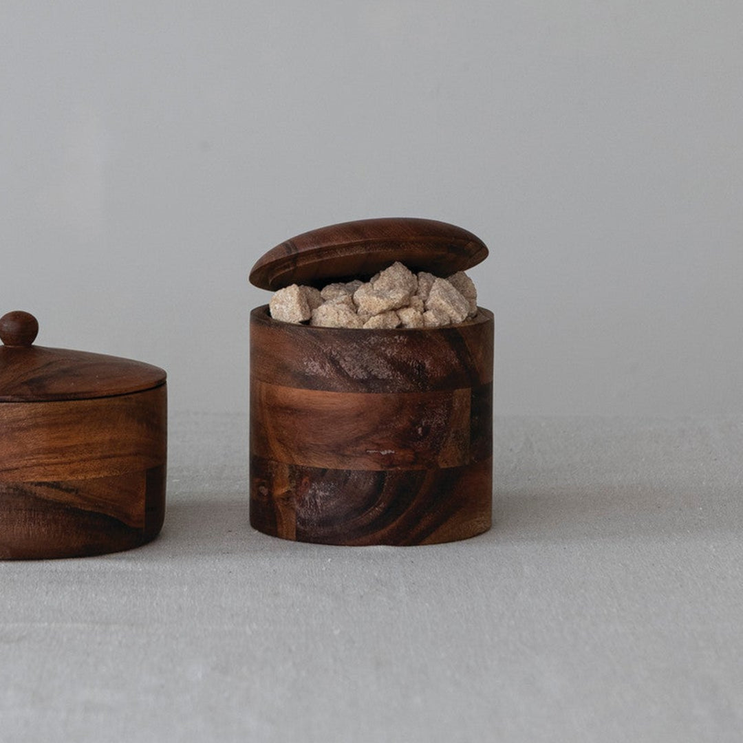 Two small round wooden containers with lids, one closed and one open showing a light-colored stone or ceramic insert, placed on a light fabric surface against a plain light gray background.
