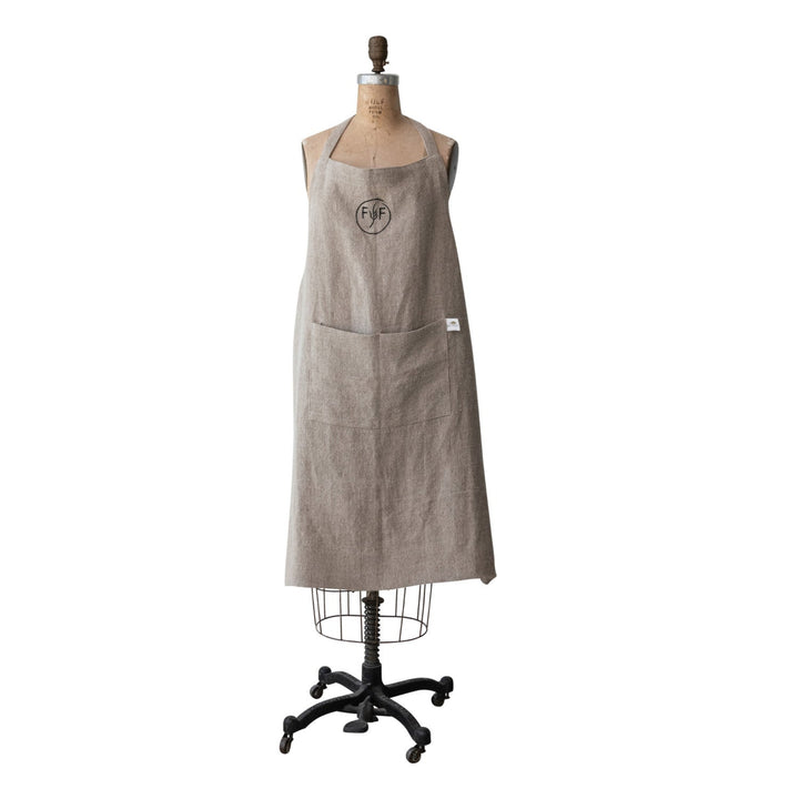 A light beige linen apron with a circular logo on the chest, displayed on a dress form mannequin stand against a solid white background.
